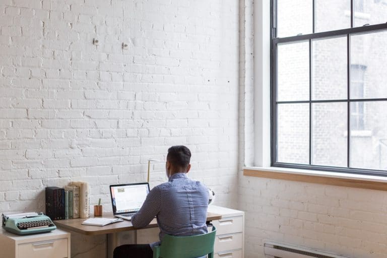 Man working on a laptop at a desk in a bright, white brick-walled home office