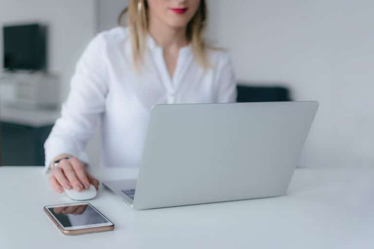 Woman in a white blouse working on a laptop with her phone on the desk beside her