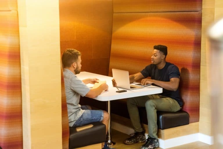 Two colleagues talk across a table in a cozy office booth, one with a laptop and notebook