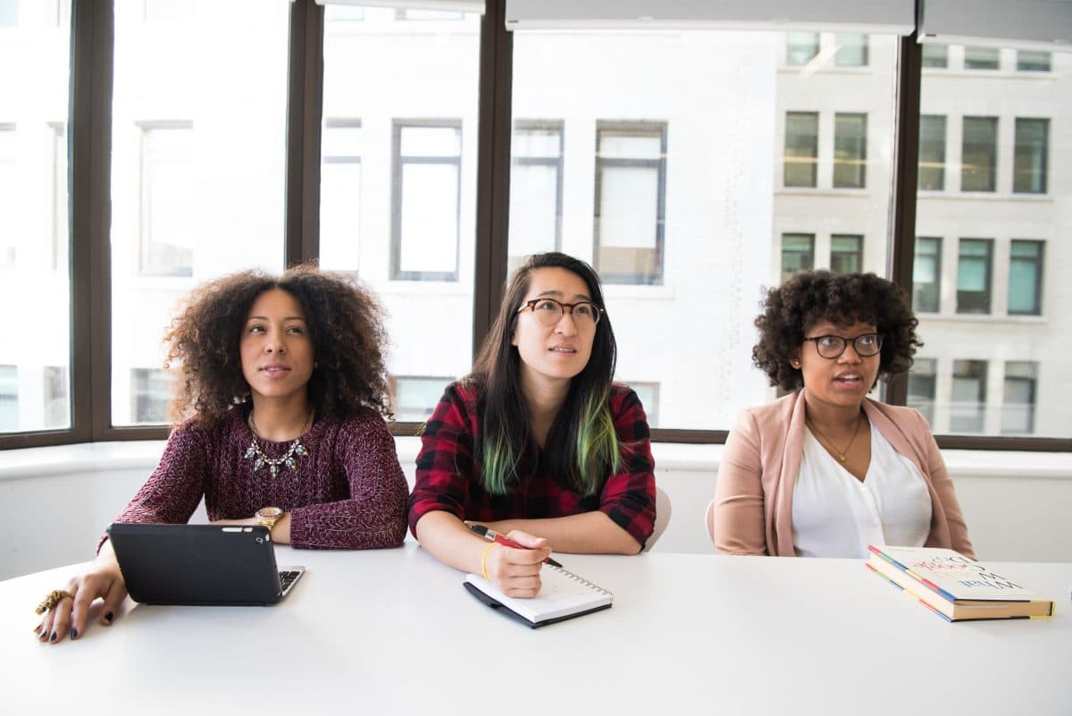 A black woman, an Asian woman, and another black woman sit behind a desk.