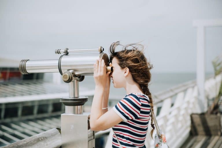 Woman looking through a coin-operated telescope on a seaside pier, scouting for talent