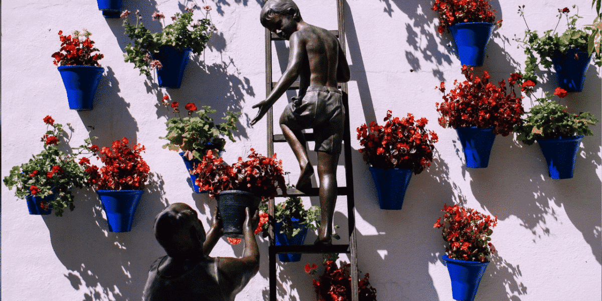 Bronze statue of a child climbing a ladder and an adult handing up a flowerpot on a wall of blue flowerpots