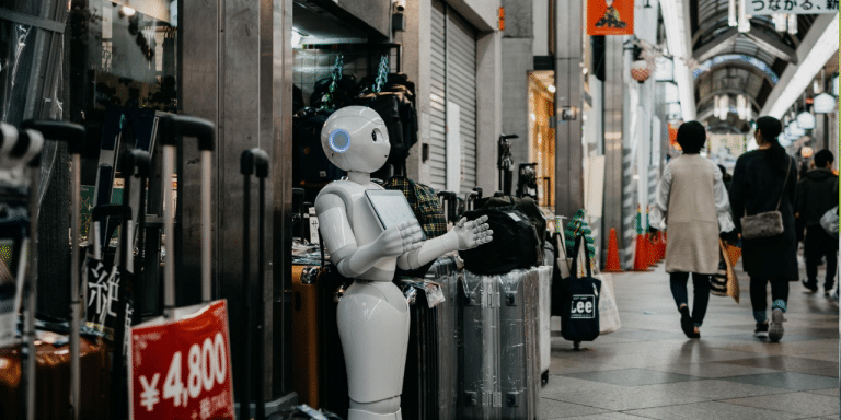 White humanoid robot holding a tablet outside a shop in a covered shopping street, as shoppers walk past