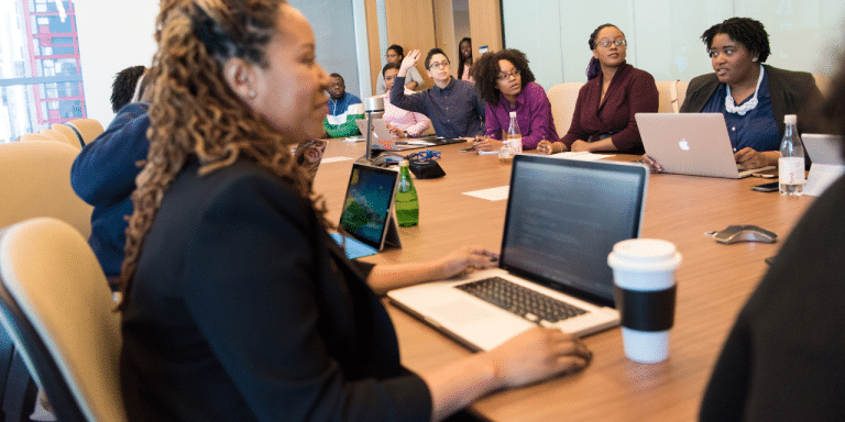 Team meeting around a conference table with laptops while a colleague raises her hand to speak