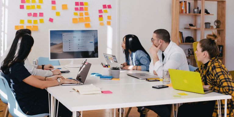 Team in a meeting looking at a screen, with a wall of sticky notes behind them
