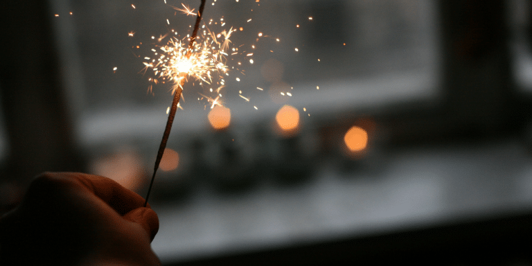 Hand holding a lit sparkler in front of a window with blurred lights