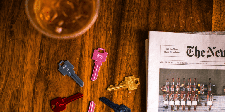 Colorful house keys scattered on a wooden table next to a newspaper and a drink