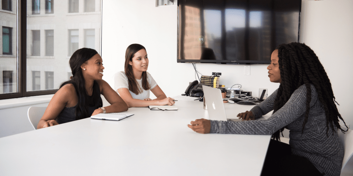 Three women sat at a table with notepads and a laptop, chatting and smiling