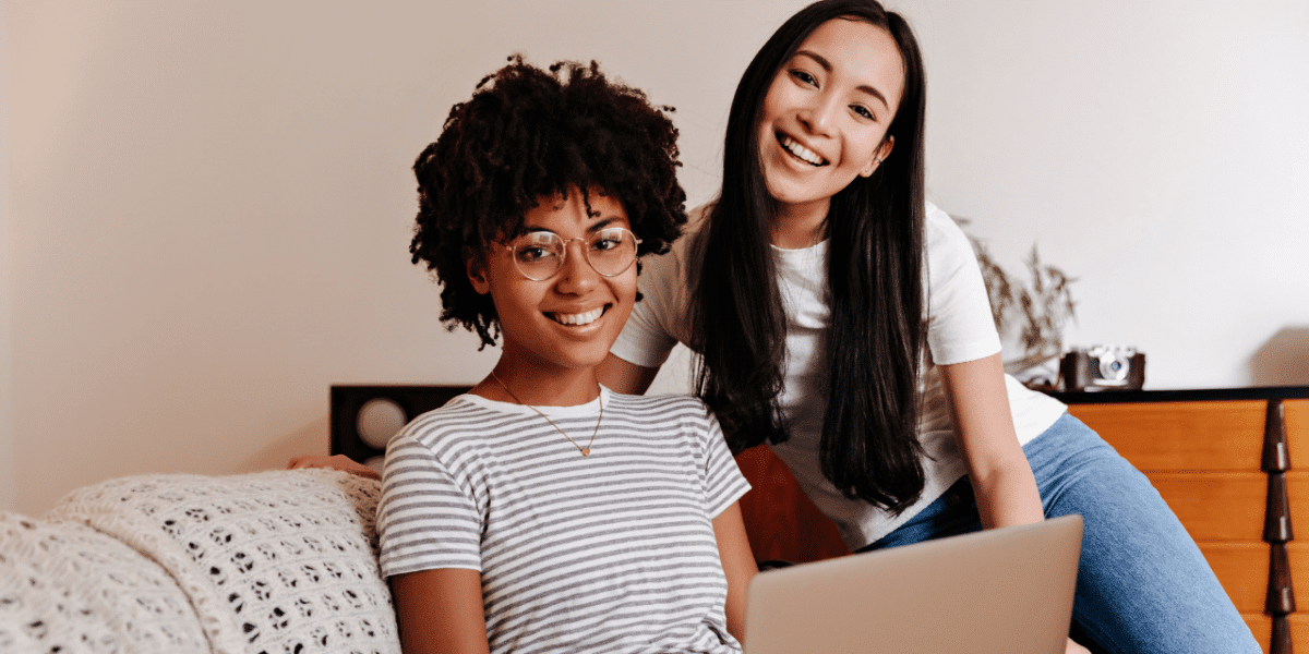 Two young women smiling at the camera with a laptop in front of them in a casual work setting