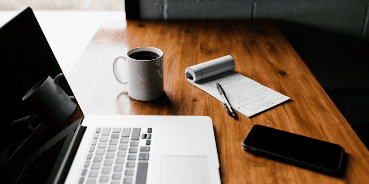 Pen and paper with a list on, next to a laptop, phone and coffee cup