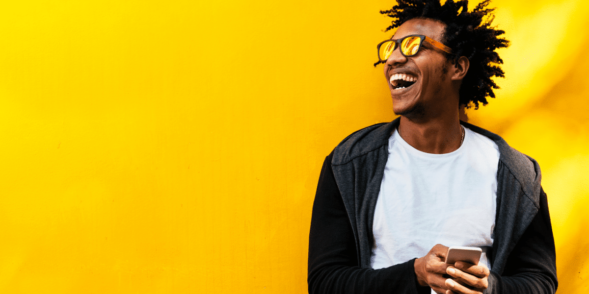 A man with sunglasses on laughing and holding his phone against a bright yellow wall