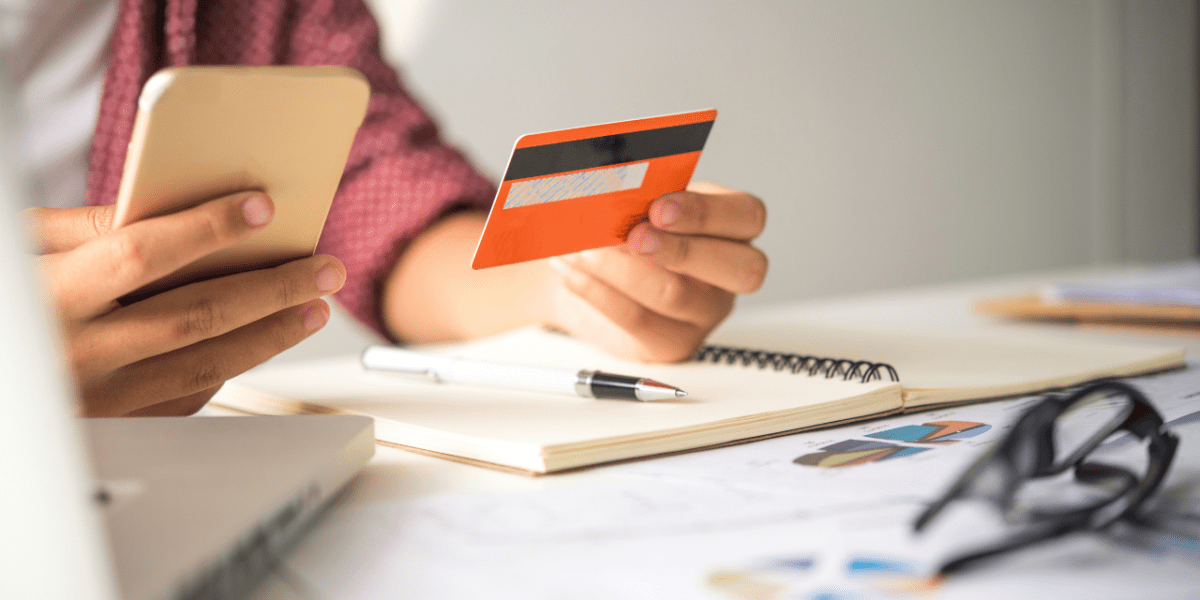 A person making a payment on their phone, holding up their credit card. They're on a busy working desk with a notebook, laptop and glasses.