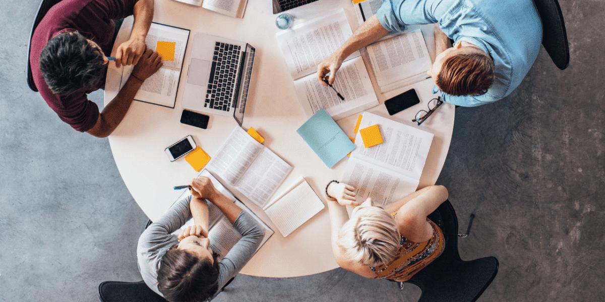 Birds-eye view of 4 people at a table covered in laptops and books, having a discussion and reading