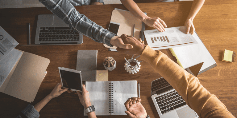 A birds-eye view of a busy desk full of open books, paper, pens, a few laptops, phones, and coffee cups. Two people are leaning over the desk to shake hands signifying a deal has been made.