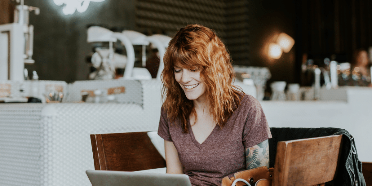 A woman with orange hair and tattoos working on her laptop in a relaxed coffee shop setting, with her bag and coat next to her. She is smiling at her laptop.