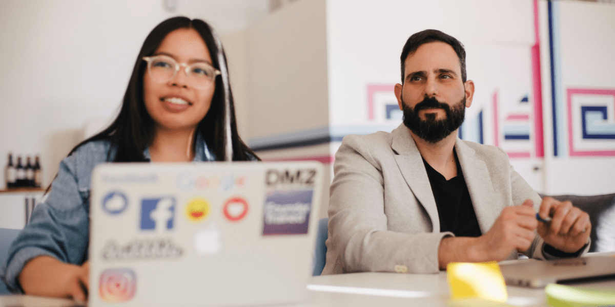 A man and a woman in a colourful, interesting workplace seated at a conference table. They're both looking ahead, past the camera, with notepads and laptops. 