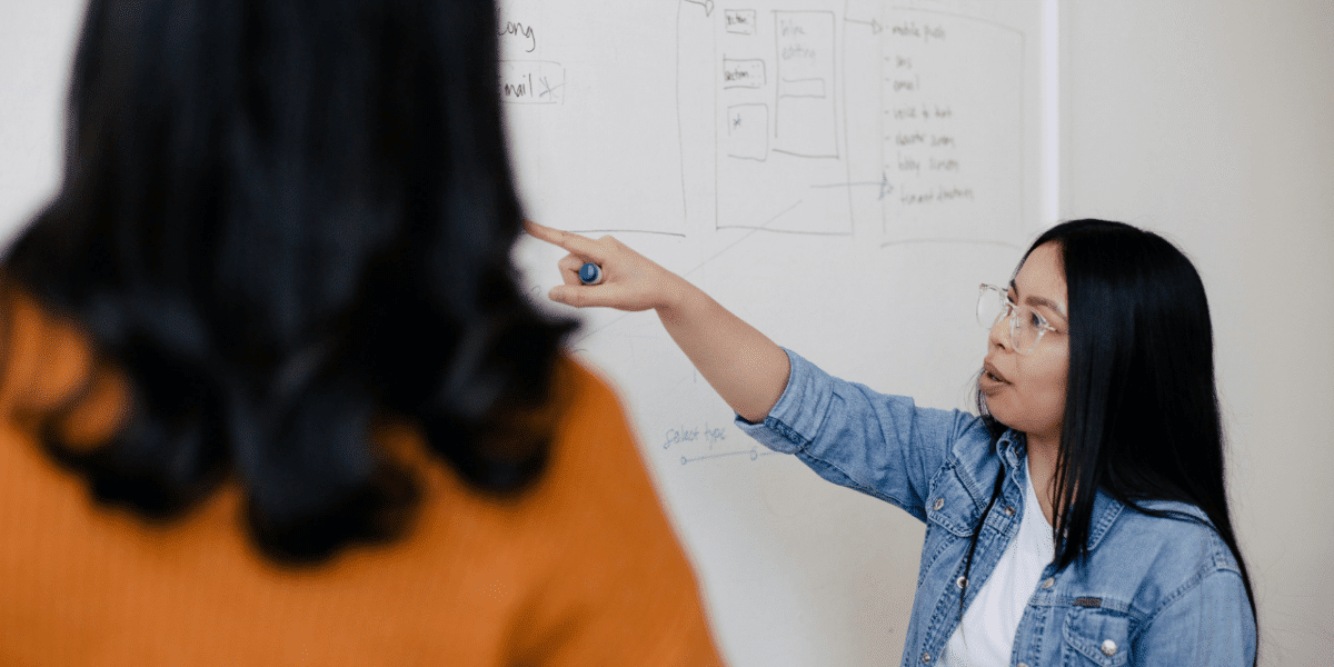 The same woman from the previous image is pointing at a whiteboard with lots of notes on. She's talking to another woman who has her back to the camera.