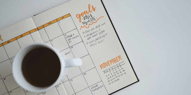 An open diary planner with a calendar spread. 'Goals this month' is written at the top of the page. A cup of coffee sits on top of the open pages