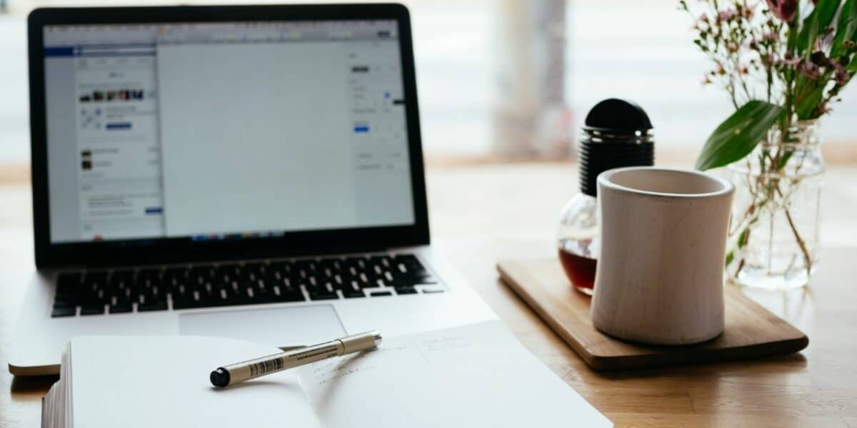 An open laptop showing an organisational program, with a notepad and pen in front of the laptop. There is also a cup of coffee and small plant on the desk.