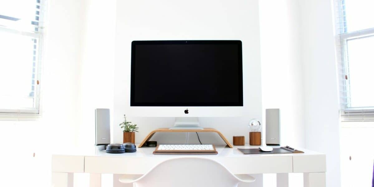 A bright, fresh home workspace. A large computer screen with two speakers either side, a keyboard and a small plant on top of a white desk with a white chair. The walls are white and there are windows either side of the desk, giving it a really bright and fresh feeling.