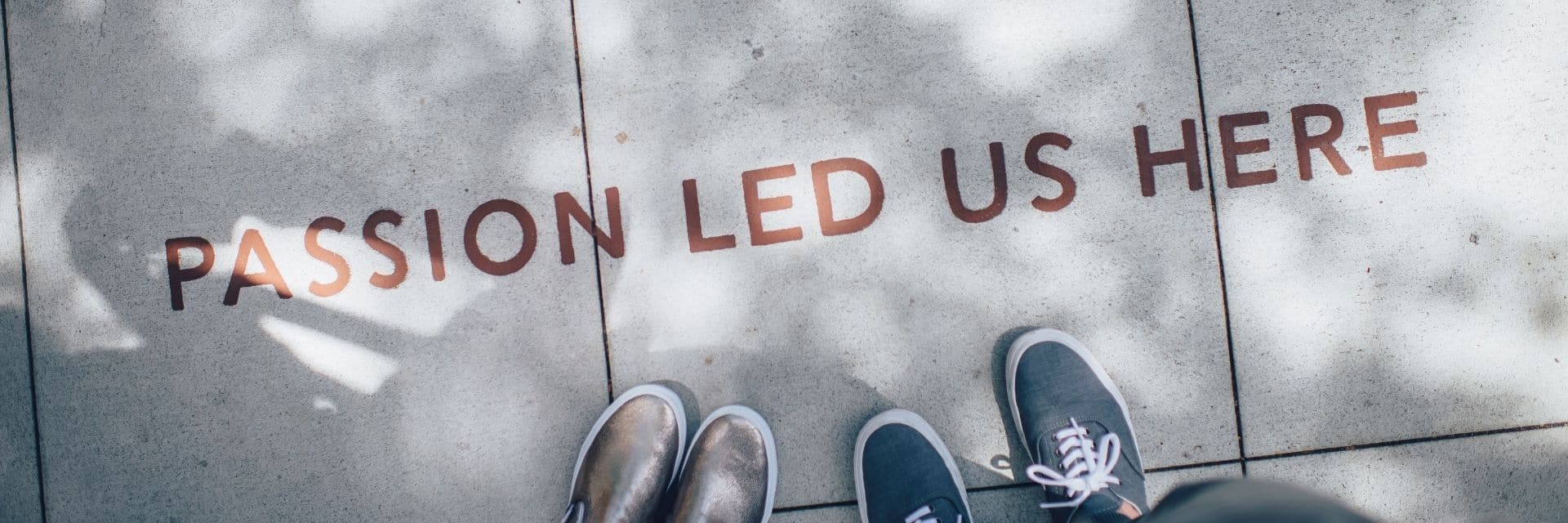 Birds-eye view of the pavement, with 2 people's shoes just in view. There's writing on the pavement reading 'PASSION LEAD US HERE' in red