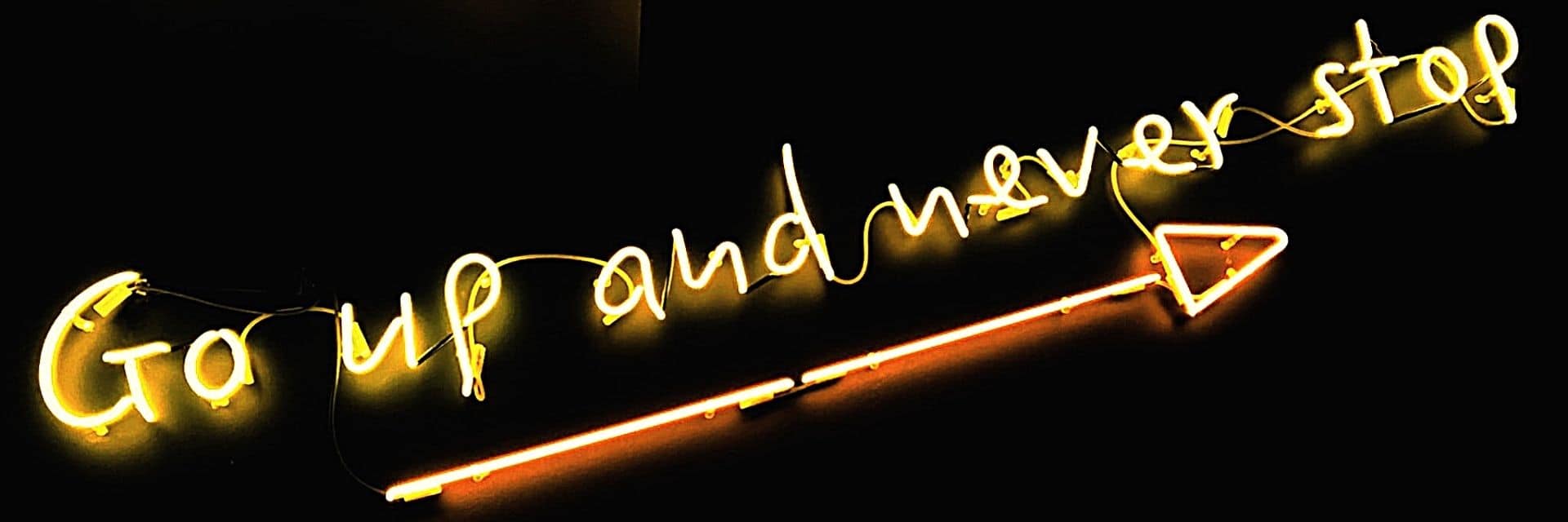 Slanted yellow neon sign reading 'go up and never stop' with an orange neon arrow pointing upwards underneath it