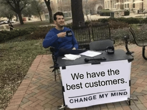 A smug-looking man sitting at an outdoor table, holding a mug. There is a sign on the table that reads: "We have the best customers. Change my mind."