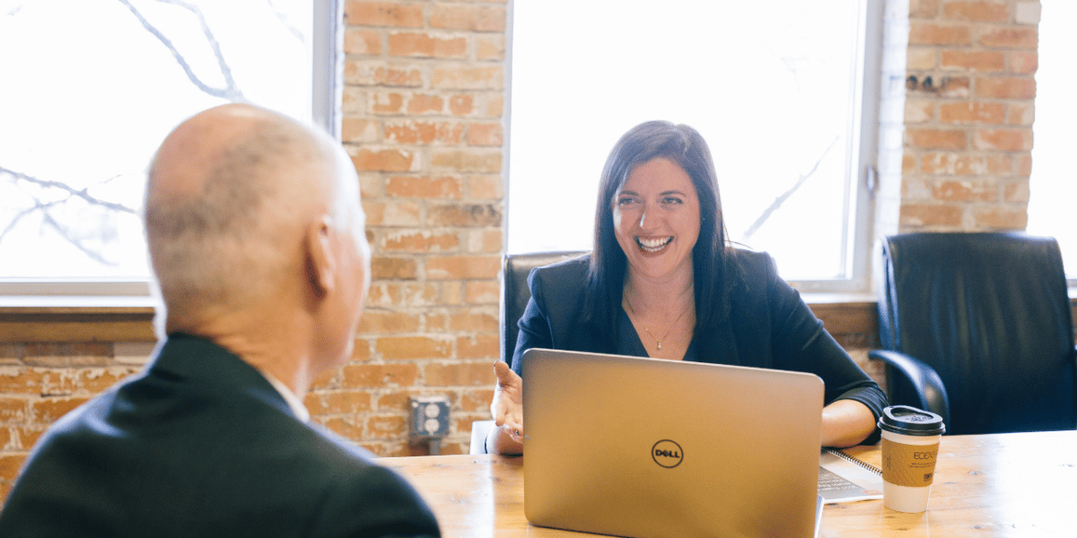 Smiling woman talking with a colleague across a table with a laptop in a brick-walled office