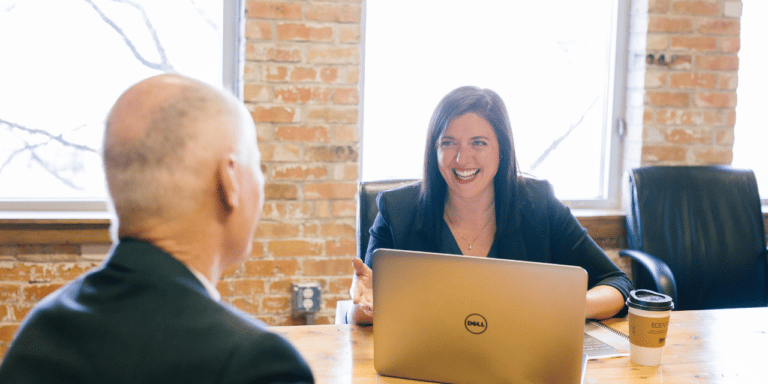 Smiling woman talking with a colleague across a table with a laptop in a brick-walled office