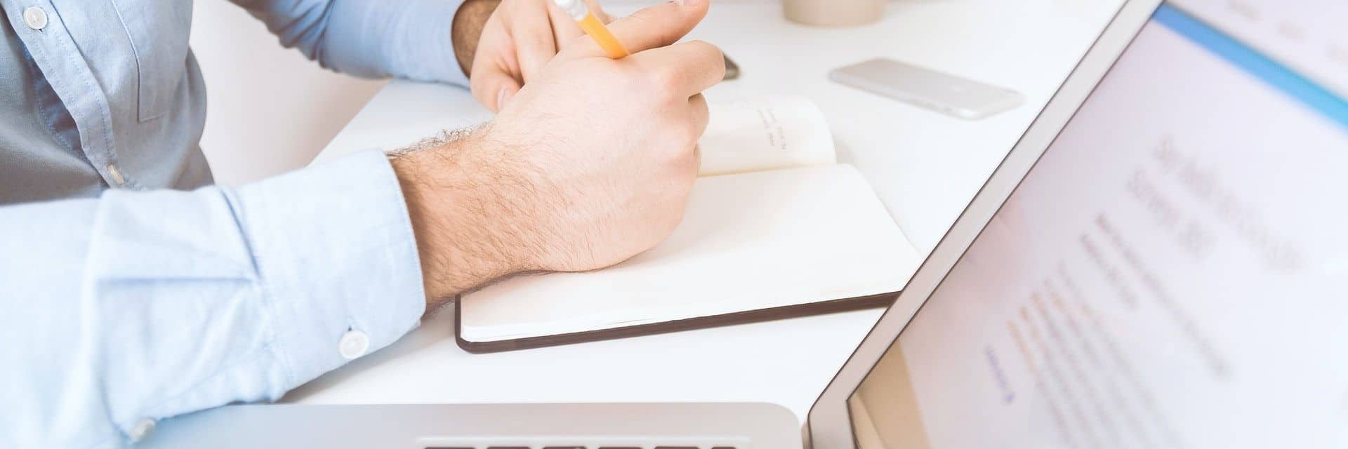 Arms resting on a table, one hand holds a pen. A notebook and laptop are visible.