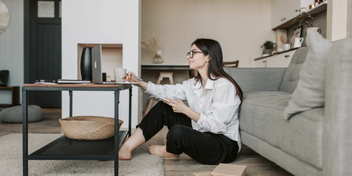 A woman sitting on her living room floor, with her laptop on the coffee table. She is surrounded by books and is leaning forward to see her laptop