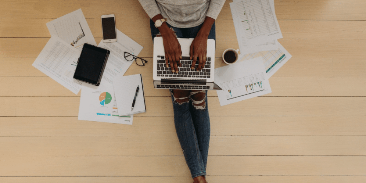 A birds-eye view of a POC sitting on the floor with their laptop on their lap. Either side of them is scattered pages, a coffee, and their phone