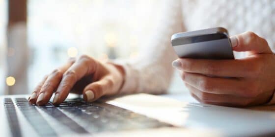 A low angle view of someone typing on a laptop with one hand, and their phone in the other hand. 