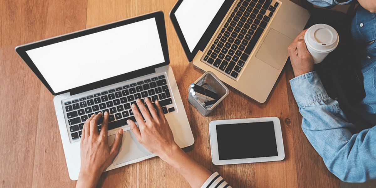 Two laptops on a desk with two people's arms in shot. There's also coffees and a tablet in view on the table.