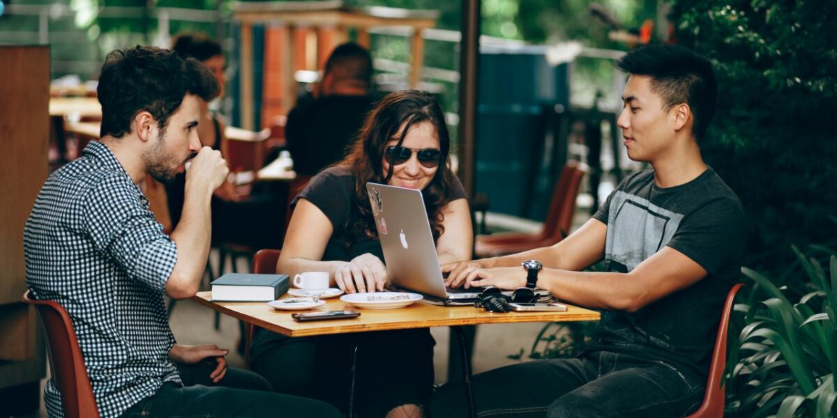 A group of three young adults at a table seated outside. The table has has notepads, coffees and a laptop on it. The three are chatting and laughing. 