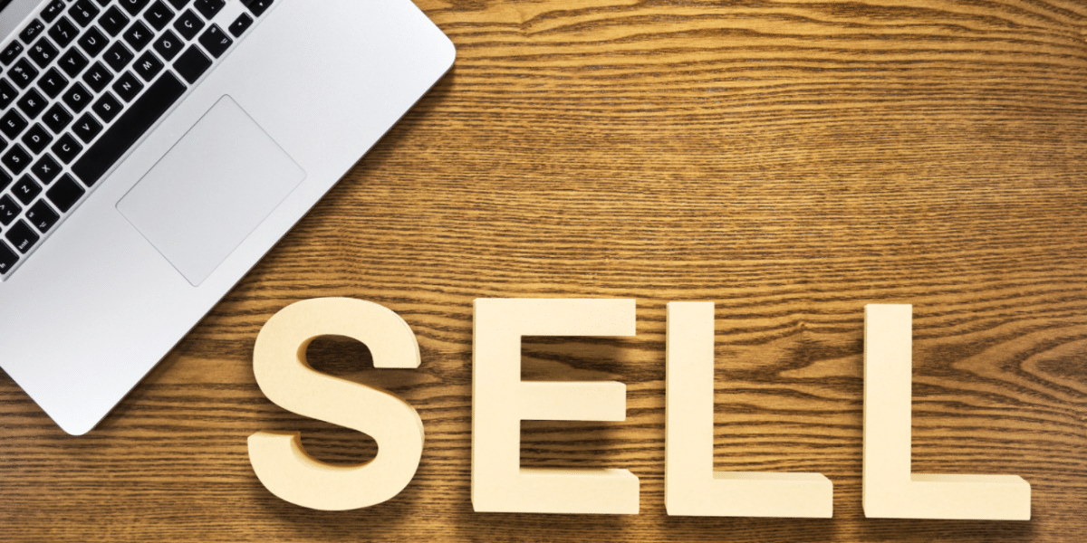 Big letters spelling out 'SELL' on a desk. In the corner of the image there is an open laptop just about in view.