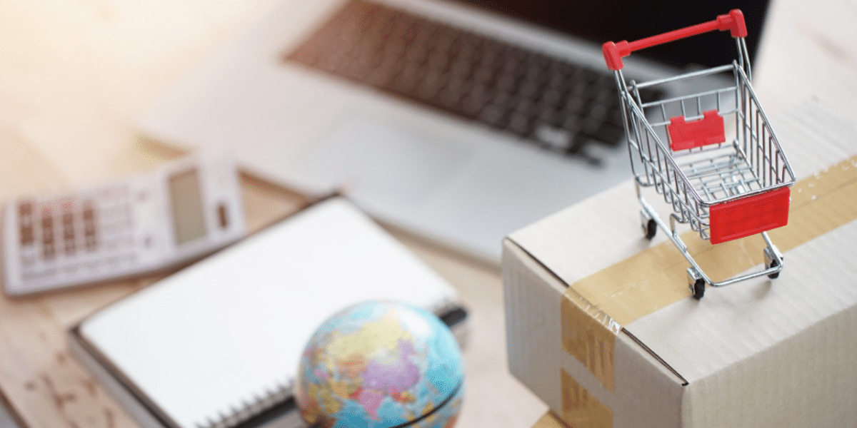 A desk showing an open laptop, a taped up box, a calculator and a mini shopping cart