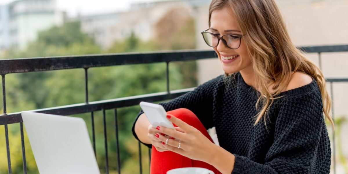 A woman sitting on a balcony smiling at her phone. Her laptop is open in front of her