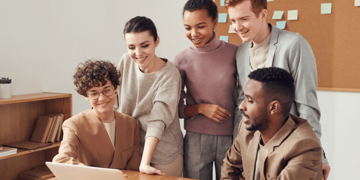 A group of 5 young people gathered around a laptop, all smiling and talking suggesting they are working together as a team