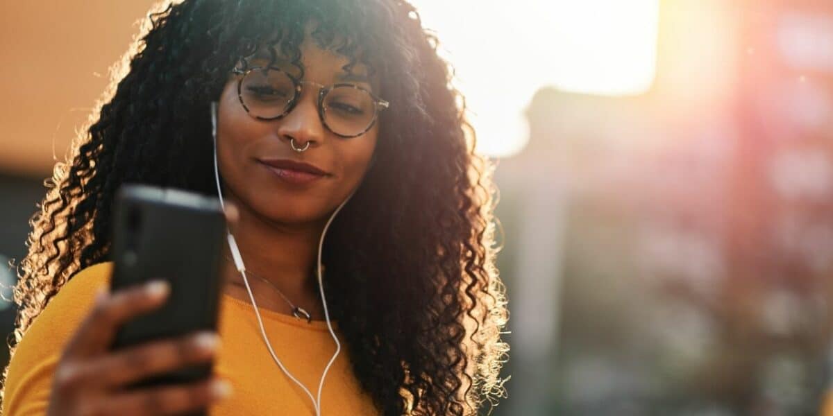 Smiling woman with earphones looking at her phone outdoors at sunset