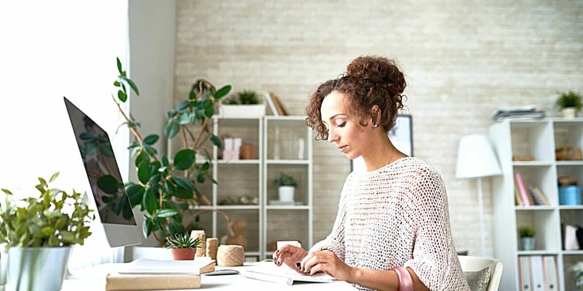 A woman looking through a notebook at her desk, with a computer screen in front of her. She's in a bright, airy office facing a window.