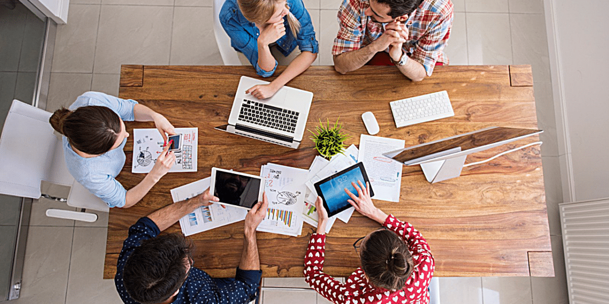 a birds-eye view of a busy table full of devices, paper, coffee. There are 5 people gathered around it all chatting and using the devices on the busy desk.