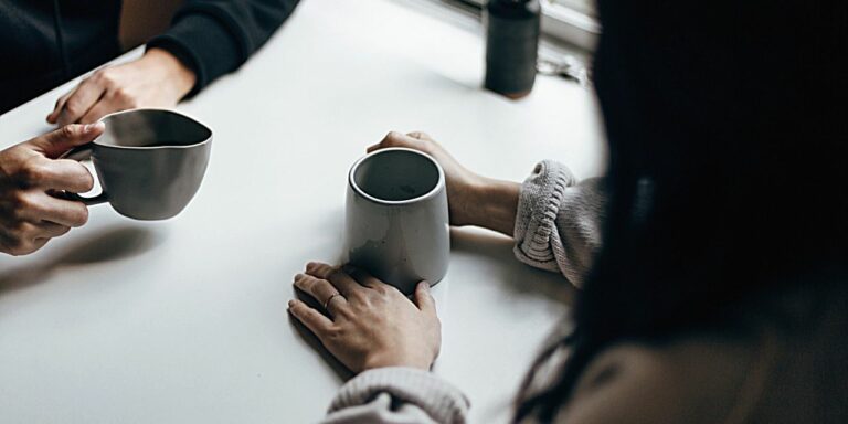 Two people opposite each other, each with a coffee cup in hand. The image only shows their arms and the cups.