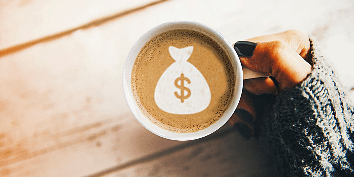 A hand holding a coffee cup. There's a bag of money shape in the coffee foam.