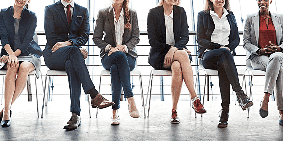 A row of people sitting on chairs, in a waiting room. Their heads are cut out of the photo, only showing them from shoulders down. Everyone is dressed in smart work attire.