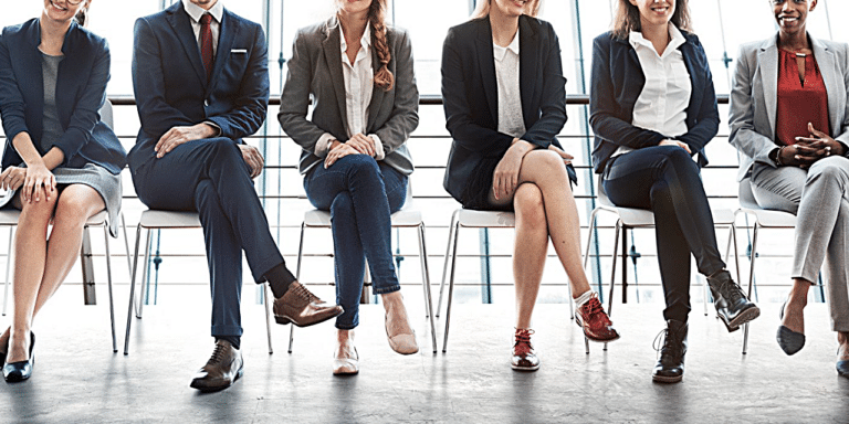 A row of people sitting on chairs, in a waiting room. Their heads are cut out of the photo, only showing them from shoulders down. Everyone is dressed in smart work attire.